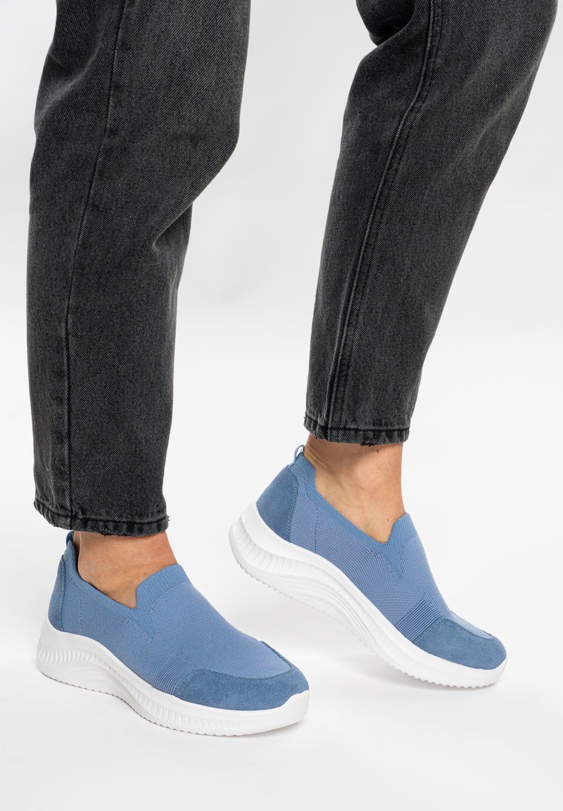Personne portant un jean noir et des baskets bleu marine à enfiler en maille avec des semelles épaisses blanches, debout sur une surface blanche.