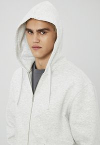 Sweat à zip gris avec un design ajusté, poches kangourou et capuche à cordon. Texture douce et apparence décontractée.