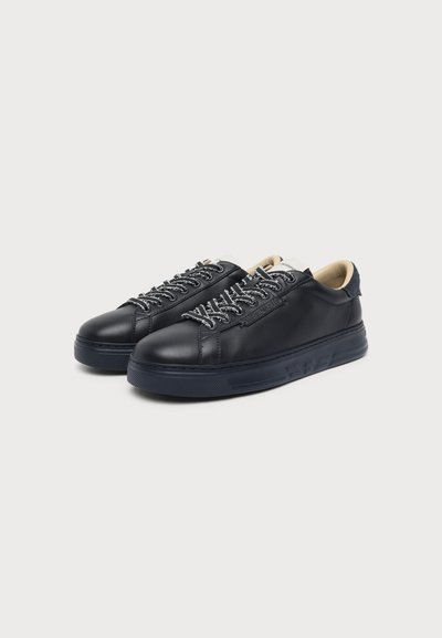 Paire de baskets basses en cuir noir avec des lacets noirs et blancs, des semelles en caoutchouc foncé, et une doublure intérieure beige sur fond blanc.