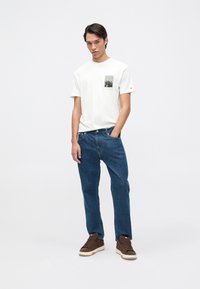 Camiseta crema con un bolsillo gráfico negro, combinada con jeans de mezclilla azul y zapatillas marrones con suelas blancas.