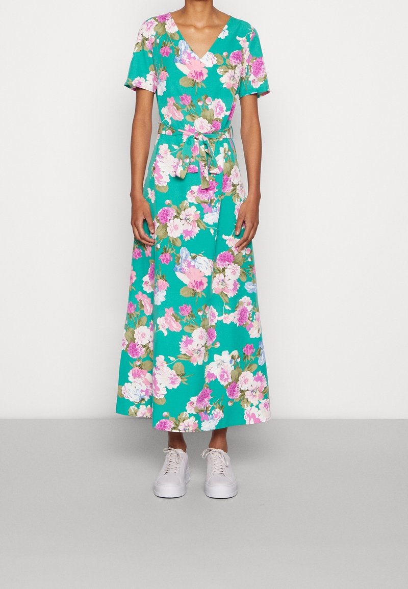 Femme portant une robe portefeuille midi fleurie bleu sarcelle à manches courtes et des baskets blanches, debout devant un fond uni.