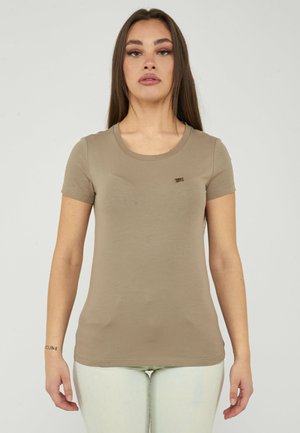 Donna con capelli castani lunghi che indossa una maglietta beige aderente a maniche corte e pantaloni chiari, in piedi davanti a uno sfondo semplice.