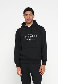 Zwarte hoodie gemaakt van zacht materiaal, met een zak aan de voorkant, een capuchon met trekkoord en een wit "Tommy Hilfiger" logo met jaartal en vlagdetail.