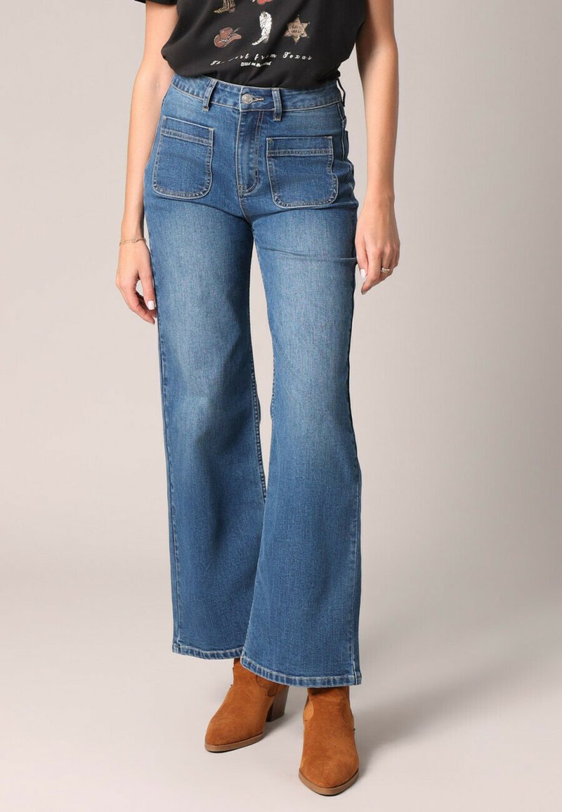 Jean en denim bleu à taille haute, évasés, avec deux poches avant. Porté avec un t-shirt graphique noir et des bottines brunes. Texture lisse.