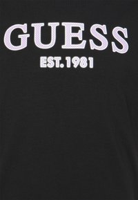Sort bomulds t-shirt med fedt, hævet hvidt "GUESS" tekst og "EST. 1981" i mindre lilla prikker. Enkel design.