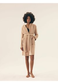 Beżowy szlafrok z kapturem, wykonany z teksturowanej tkaniny, z długimi rękawami, paskiem w talii i dwiema przednimi kieszeniami. Rękaw kończy się powyżej kolana.