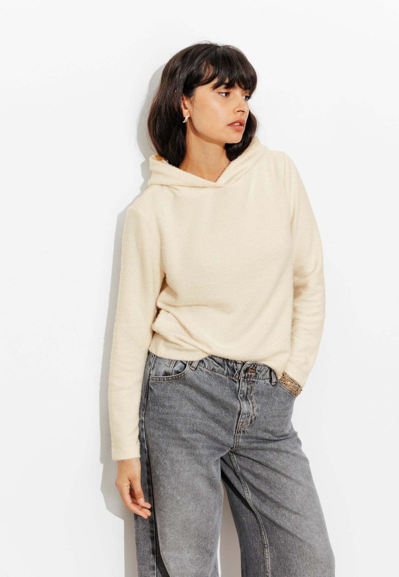 Sweatshirt à capuche en créme, fabriqué en tissu texturé, associé à un jean en denim gris taille haute. Présente une coupe décontractée et des poches avant.