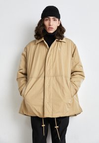 Beige gewatteerde jas van lichtgewicht, gestructureerde stof, met knoopsluiting en twee zijzakken. Zwarte geribbelde beanie gedragen.