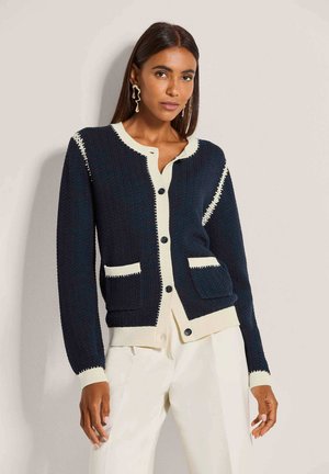 Vrouw draagt een donkerblauwe cardigan met crèmekleurige bies en knopen, gecombineerd met witte broek, staand tegen een lichtbeige muur.