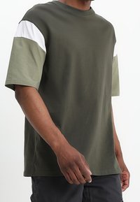Camiseta de algodón en verde oliva con mangas cortas, que presenta paneles en verde claro y blanco en los hombros. Cuello redondo clásico, ajuste relajado.