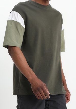 T-shirt in cotone verde oliva con maniche corte, caratterizzato da pannelli verdi chiari e bianchi sulle spalle. Collo rotondo classico, vestibilità comoda.