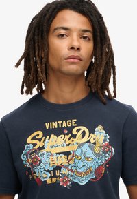 Camiseta de algodón azul marino con un diseño gráfico colorido que presenta un rostro azul, flores rojas y letras doradas. Con mangas cortas y cuello redondo.