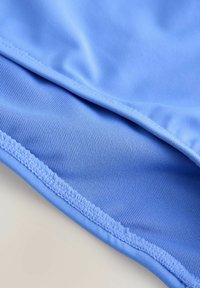 Lyseblåt stof med glat tekstur og fint ribmønster. Buet kant med synlige stikninger, der viser en ren finish.