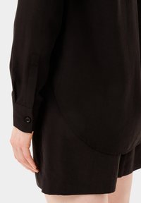 Camicia nera a maniche lunghe, con orlo arrotondato e polsini con bottoni. Il tessuto ha un aspetto liscio, leggermente testurizzato.