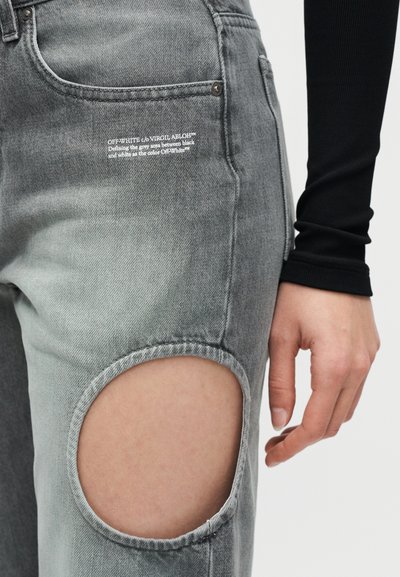 Γκρι τζιν denim με μεγάλο άνοιγμα στο γόνατο, λευκό τυπωμένο κείμενο κοντά στην τσέπη και ένα χέρι που ακουμπάει δίπλα στο μηρό φορώντας μαύρο μπλουζάκι με μακριά μανίκια.