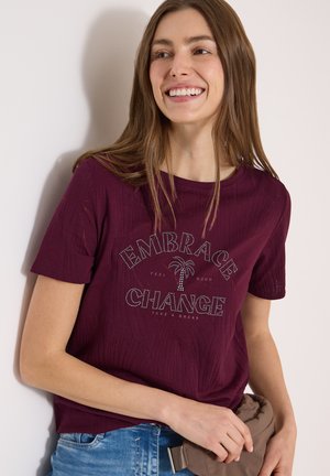 Lächelnde Frau mit langen braunen Haaren, die ein dunkelrotes Shirt mit der Aufschrift „Embrace Change“ und einer Palmen-Grafik trägt und einen braunen Gegenstand in der Taille hält.