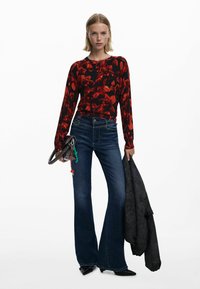 Piros virágmintás, hosszú ujjú blúz, amely deréknál tökéletesen illeszkedik, sötétkék, bővülő farmernadrággal párosítva. Fekete kézitáskát és szürke dzsekit visel.