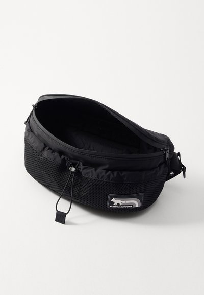Sac banane noir avec un tissu supérieur lisse et un fond en maille texturée, muni d'une fermeture éclair et d'un patch logo pour une identification facile.
