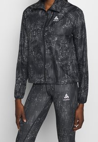 Chaqueta atlética negra con cierre de cremallera, que presenta un patrón de puntos y un logo en el lado izquierdo del pecho. Combinada con leggings a juego.