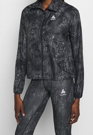 Musta urheilutekotakki, jossa on vetoketjukiinnitys, jossa on pistekuvio ja logo vasemmassa rinnassa. Yhdistetty vastaavaan leggingseihin.