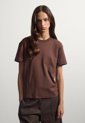 Donna con capelli castani lunghi che indossa una camicia marrone a maniche corte e pantaloni scuri, in piedi davanti a uno sfondo chiaro e uniforme.