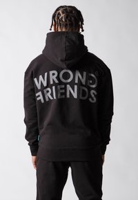 Felpa nera con cappuccio e la scritta "WRONG FRIENDS" stampata in grandi lettere grigie sulla schiena. Realizzata in un tessuto morbido, con polsini e bordo a coste.