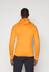 Πορτοκαλί μακρυμάνικη μπλούζα με κουκούλα και εφαρμοστή σχεδίαση. Λείο ύφασμα και αντίθετες σκούρες ραφές. Φορεμένη πάνω από σκούρα παντελόνια.