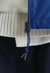 Chaqueta azul con cremallera y cuello de punto en color crema con acanalado, presenta un cordón azul anudado con puntas metálicas y una capa blanca por debajo.