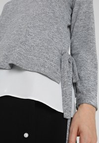 Szary sweter z dzianiny z detalem związania po boku, nałożony na białą bluzkę. Miękka faktura, casualowy krój i proste wykończenie, widoczne czarne spodnie poniżej.