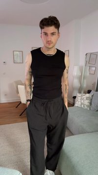 Black sleeveless shirt, loose black pinstriped pants, white sneakers, and layered necklace; standing in a modern indoor setting.