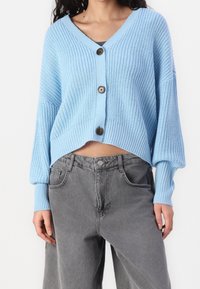 Jasnoniebieski dziergany kardigan z dekoltem w serek, zapinany z przodu na guziki, z prążkowanymi mankietami, zestawiony z szarymi dżinsami z wysokim stanem. Tkanina wydaje się mieć teksturę.