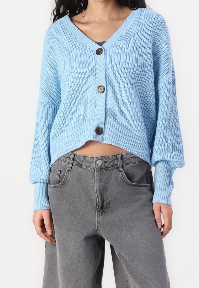 Jasnoniebieski dziergany kardigan z dekoltem w serek, zapinany z przodu na guziki, z prążkowanymi mankietami, zestawiony z szarymi dżinsami z wysokim stanem. Tkanina wydaje się mieć teksturę.
