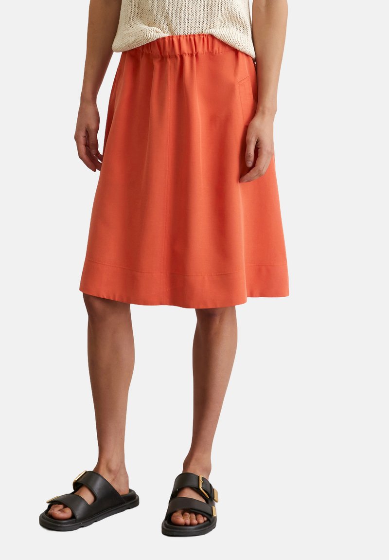 Orange knee-length skirt with an elastic waistband and a smooth texture, paired with black leather slide sandals featuring gold buckles.