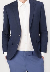 Mann in einem navy Blazer, einem weißen Hemd mit Knöpfen und blauen Hosen mit den Händen in den Taschen vor einem schlichten Hintergrund.