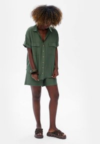 Zielona, krótko rękawowa sukienka koszulowa z zapięciem na guziki, dwoma kieszeniami na piersi i dopasowanymi szortami. Gładka tekstura i luźny fason, połączone z brązowymi sandałami.