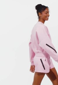 Light pink sweatshirt with dropped shoulders and matching shorts, featuring black zipper accents on the pockets. Smooth texture.