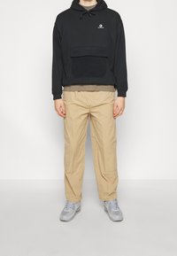 Persona che indossa un hoodie nero dei Converse, pantaloni beige e scarpe da ginnastica grigie, in piedi contro uno sfondo bianco semplice.