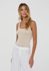 Body beige aderente con spalline larghe, scollatura quadrata e texture morbida, abbinato a pantaloni bianchi a vita alta.