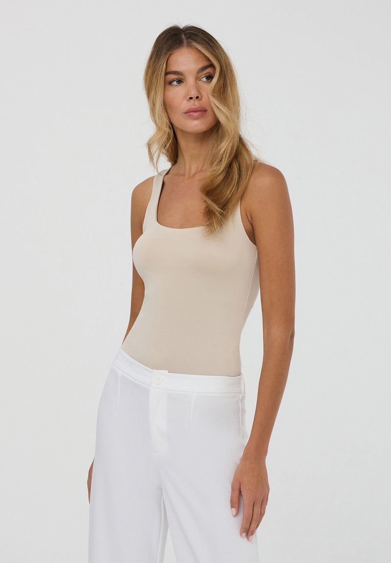 Body beige aderente con spalline larghe, scollatura quadrata e texture morbida, abbinato a pantaloni bianchi a vita alta.