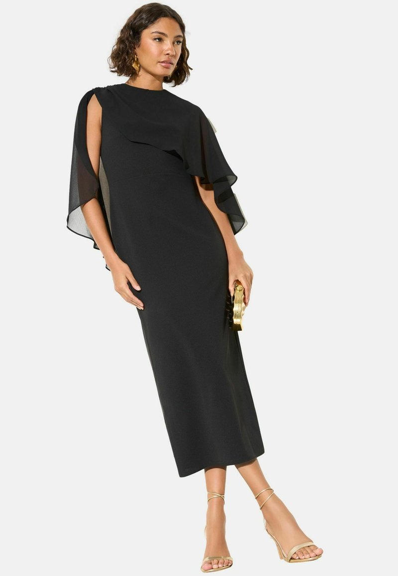 Frau trägt ein schwarzes Midikleid mit durchsichtigen Cape-Ärmeln, hält eine goldene Clutch und trägt goldene Riemchenabsätze.