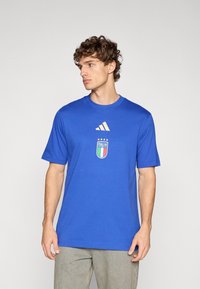 Tricou albastru din bumbac cu mâneci scurte, prezintă un logo adidas auriu și emblema echipei naționale a Italiei cu accente colorate în partea din față.