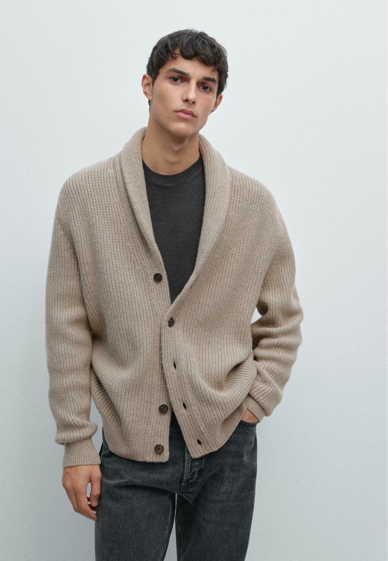 Beige Strickjacke mit Schalkragen, geripptem Textur und Holzknöpfen, kombiniert mit einem dunkelgrauen Rundhalshemd und dunklen Jeans.