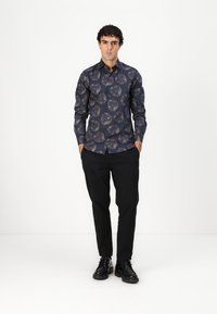 Langermet skjorte med knapper i marineblått, med et blomsterdesign i nyanser av lys brun. Stilt med svarte bukser og svarte sko.