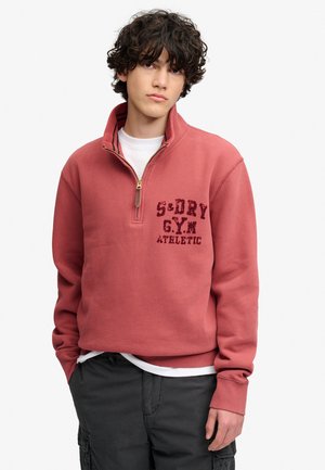 Jong persoon met krullend haar, draagt een rode halve-ritssluiting trui met de tekst "S-DRY G.Y.M ATHLETIC" en donkere cargobroek, staand tegen een witte achtergrond.