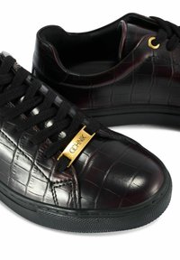 Czarne niskie trampki z teksturą krokodyla, czarne sznurowadła, złota oczko i złota zawieszka ze słowem "QCHNIK" na białym tle.
