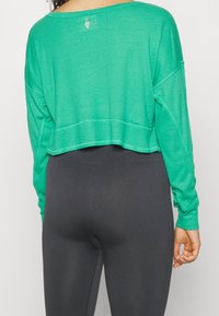 Top corto de manga larga verde hecho de una tela texturizada, con un escote ancho y un ajuste holgado, combinado con leggins gris oscuro.