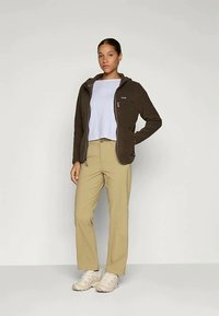 Chaqueta de forro polar marrón con cuello alto, camisa de color azul claro, pantalones beige y zapatillas blancas. Presenta un bolsillo con cremallera en la chaqueta.