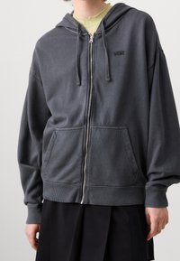 Grey zip-up hoodie made of soft fabric, featuring a hood, kangaroo pockets, and ribbed cuffs and hem; embroidered logo on the chest.