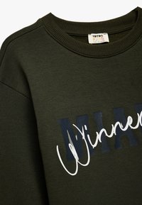 Ciemnozielona bluza z ribbowanym dekoltem, z wyrazistym napisem "Winners" w granatowym i białym kolorze. Miękki materiał i standardowy krój.