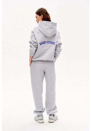 Kobieta stoi tyłem do kamery, ubrana w szarą bluzę z kapturem z napisem „NAOKO STUDIO” na plecach, dopasowane spodnie dresowe i białe trampki.
